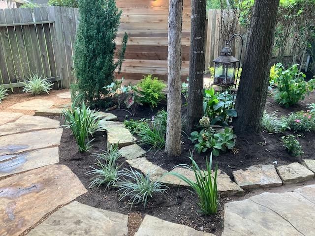 A stone path winds through a garden with trees, plants, and wooden steps in the background.