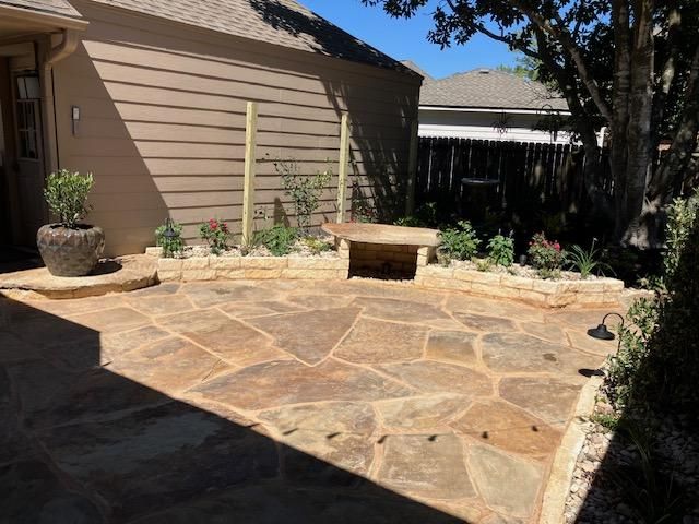 Patio with flagstone flooring, a stone bench, flowerbeds, and a tan building. Sunny outdoor setting.