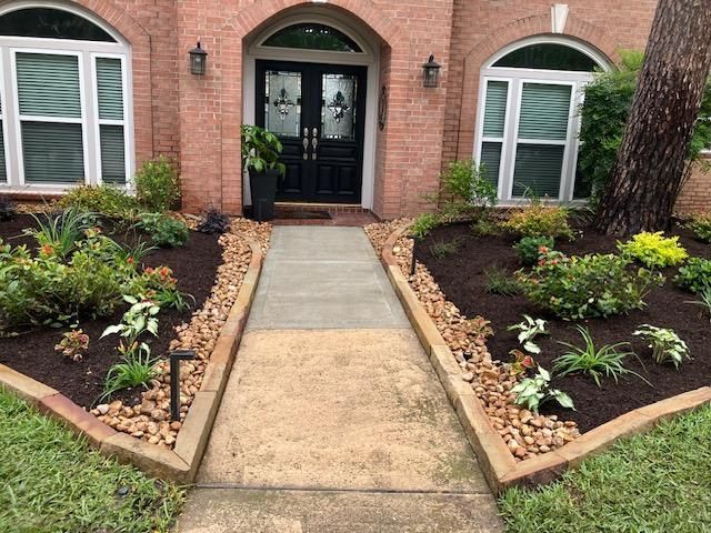Brick home exterior with pathway to black double doors, flanked by landscaped flower beds and decorative stones.