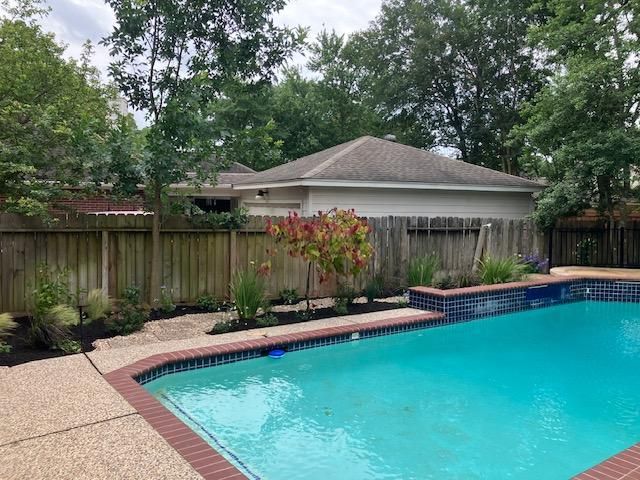 Backyard pool with a house and fence in the background, surrounded by trees and landscaping.