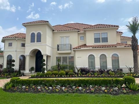 Beige stucco mansion with a red tile roof and well-manicured landscaping under a blue sky.