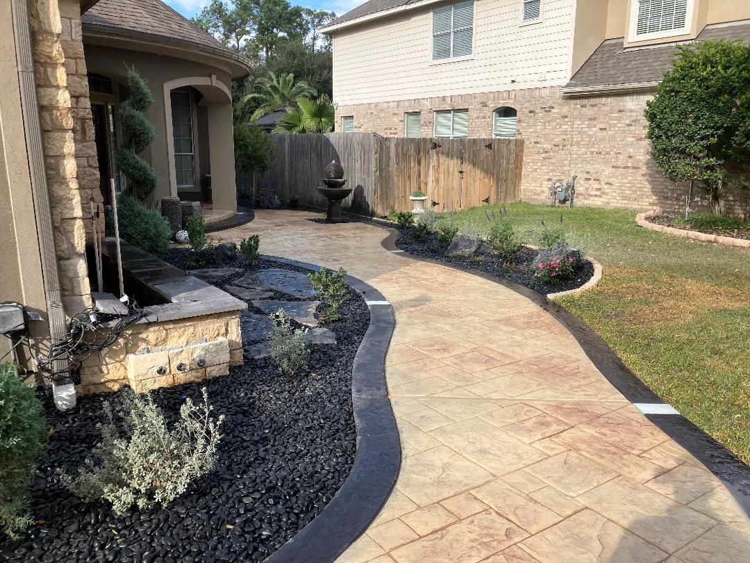 Curving stamped concrete walkway with dark border, landscaped beds, and a fountain in front of a house.