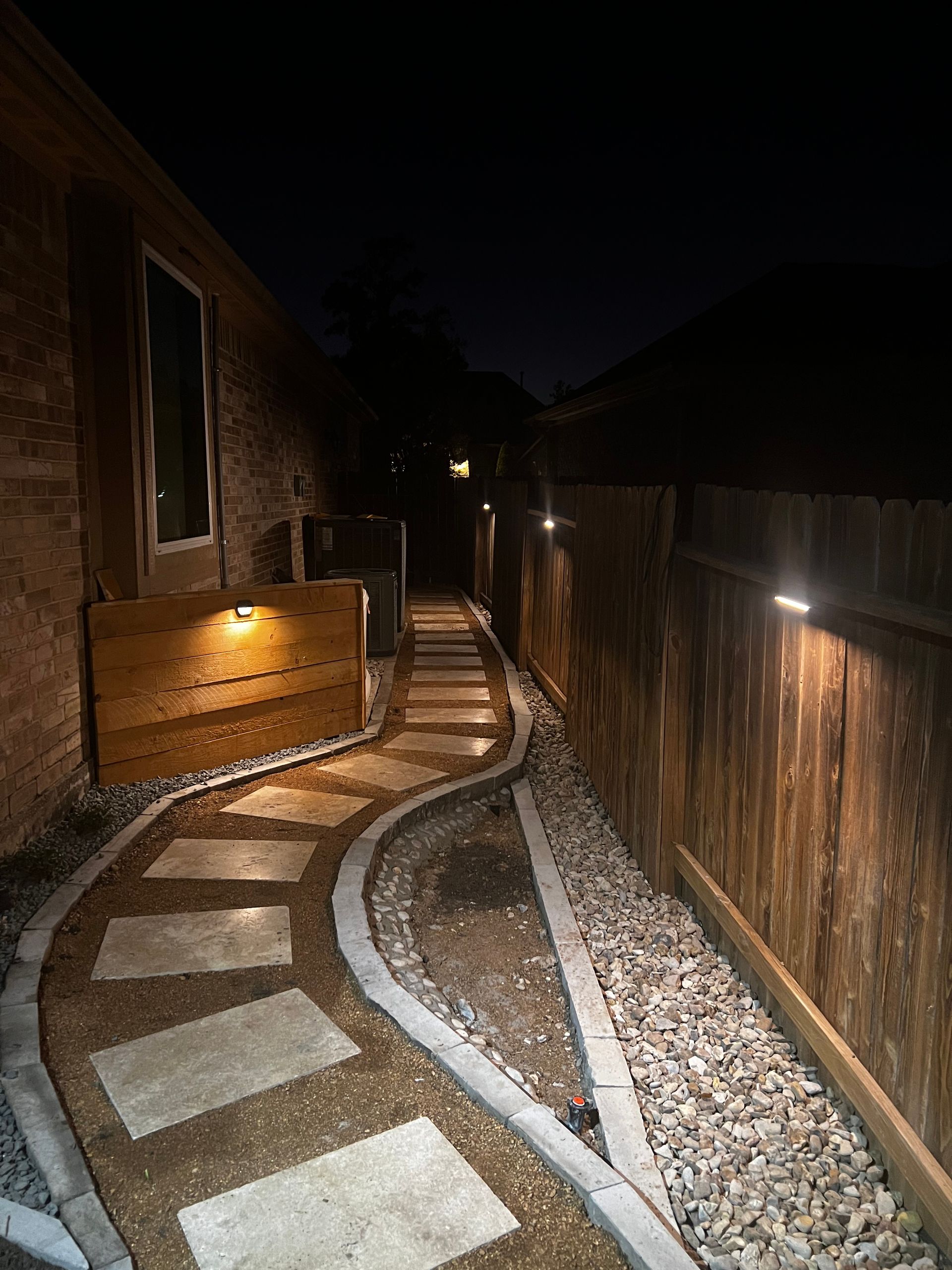 A nighttime outdoor walkway illuminated by lights on a wooden fence, leading towards the darkness.