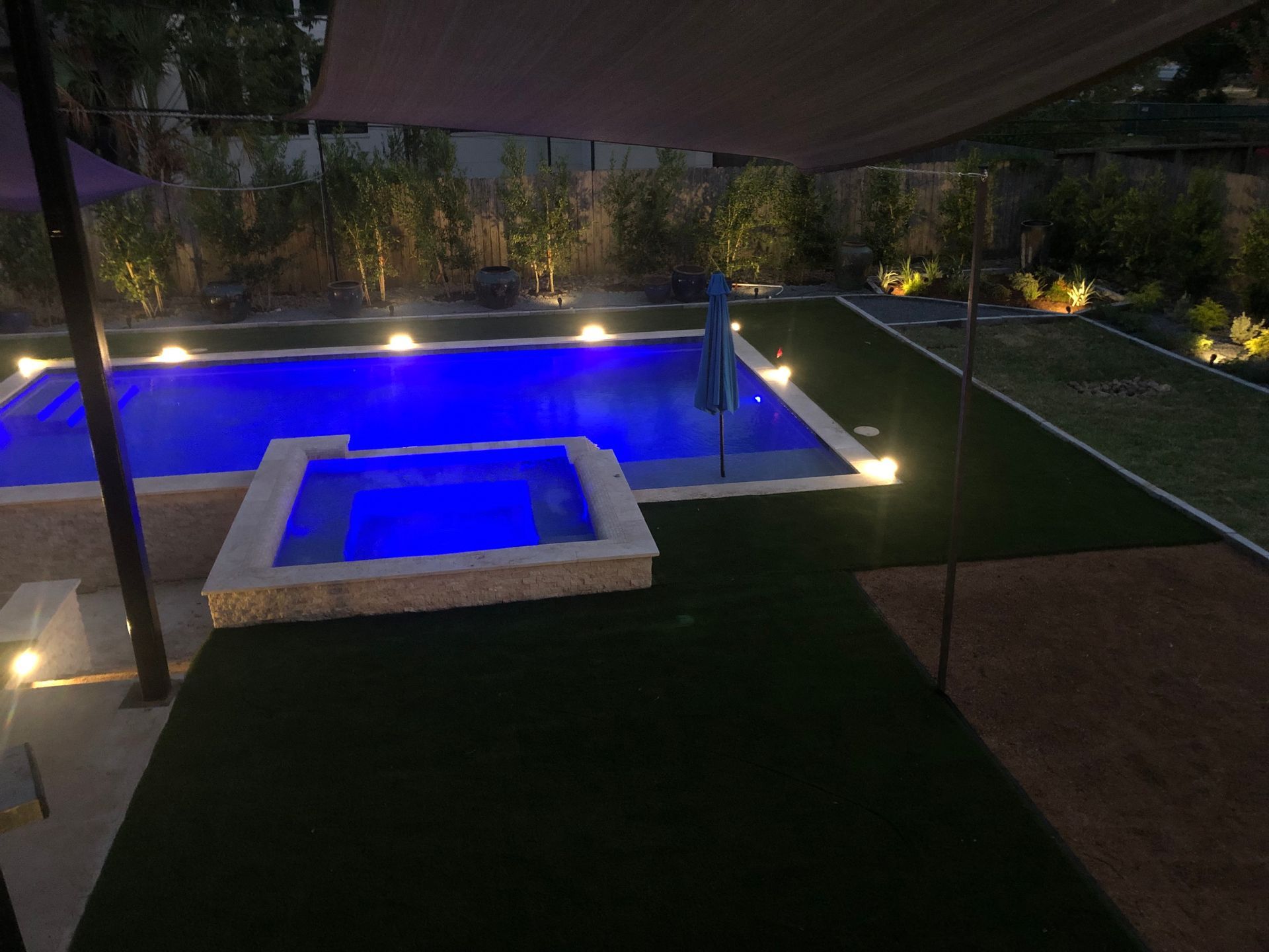 Night view of a backyard with a lit pool, hot tub, and lawn. Artificial turf and lights enhance the ambiance.