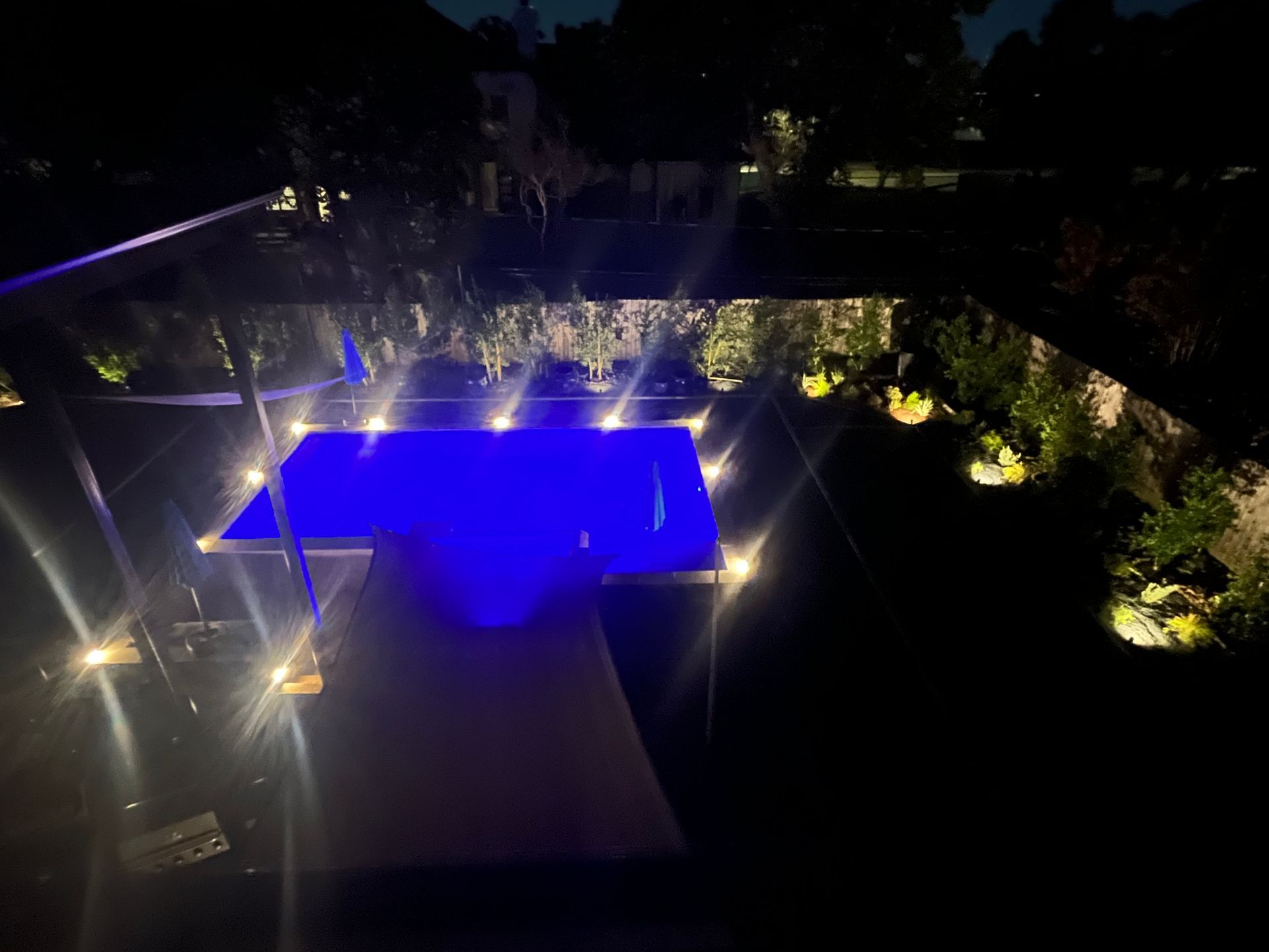 Night view of a rectangular pool with blue lights, surrounded by illuminated landscape and trees.