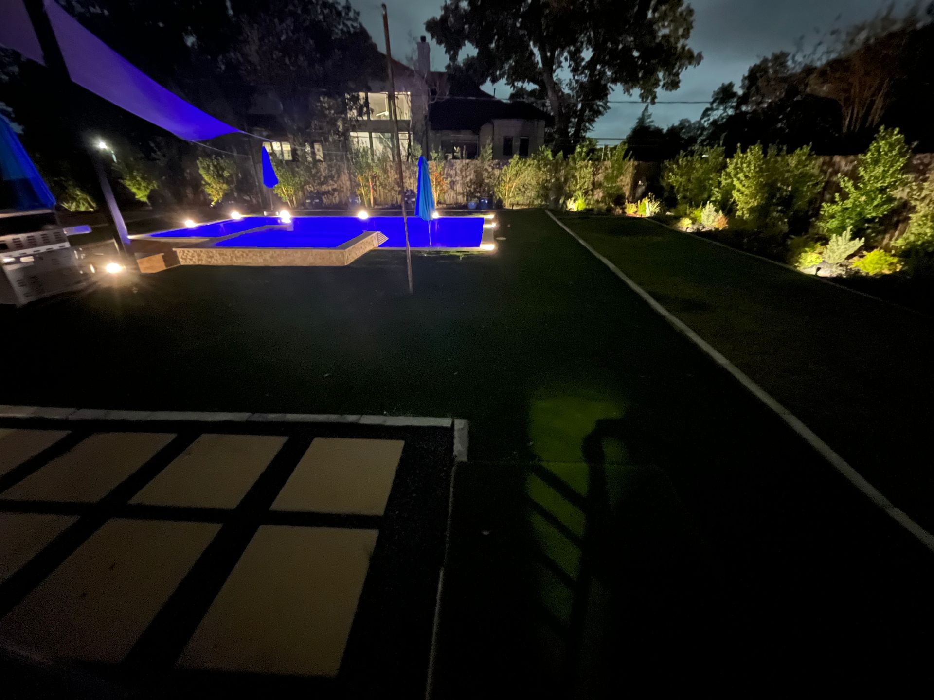 Nighttime backyard scene with a blue-lit pool and manicured lawn. Illuminated landscaping and large house in the background.
