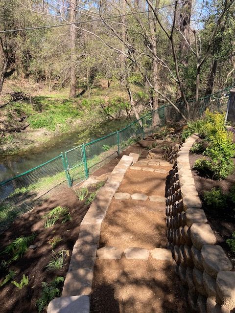 Stone steps lead down to a green chain link fence along a waterway. Lush greenery and trees surround.