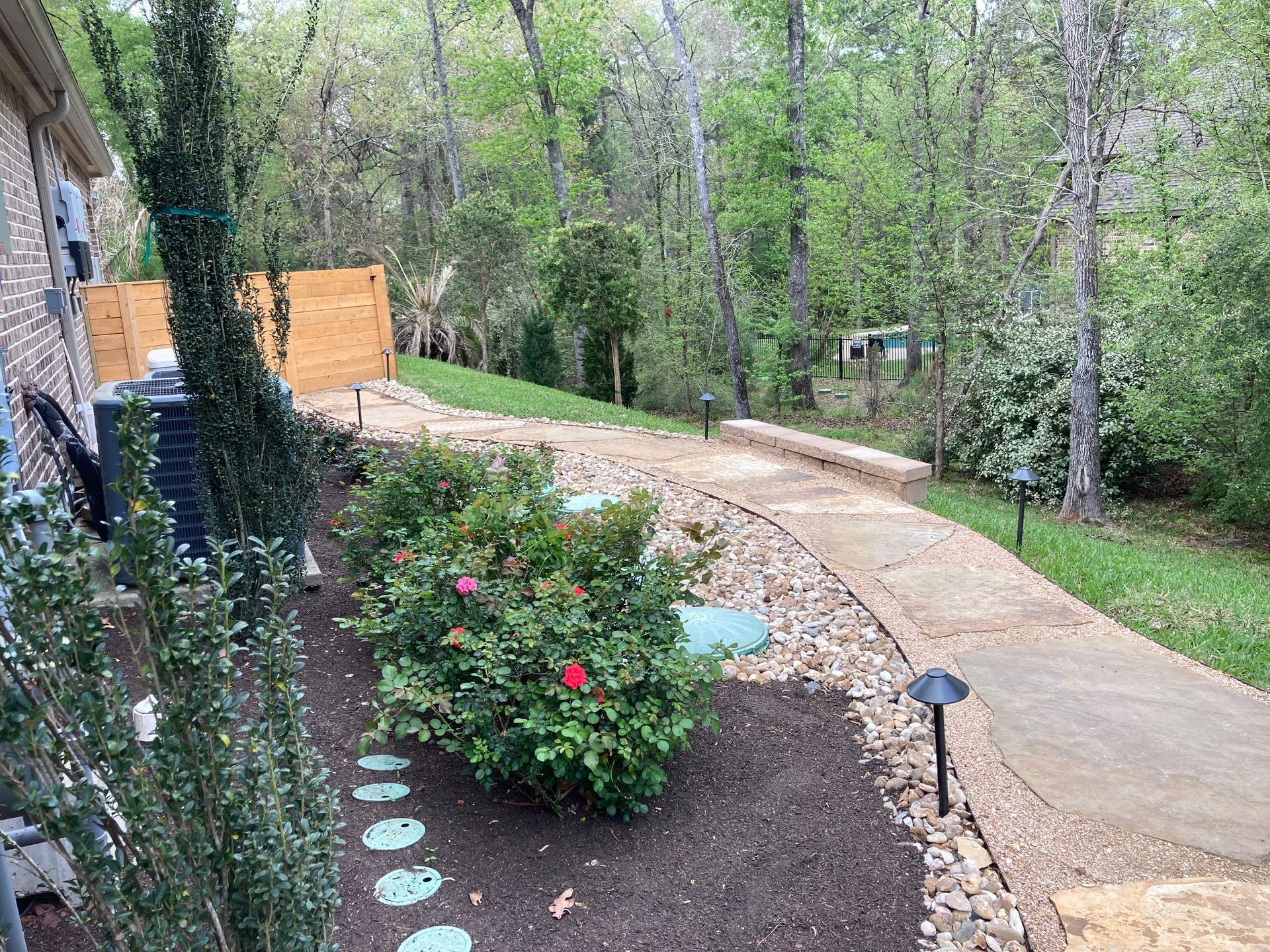 Pathway in a backyard, with landscaping and lights, leading into a wooded area.