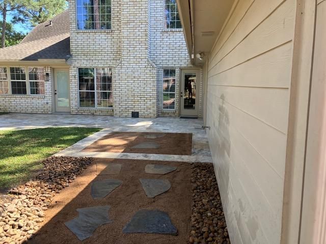 Backyard with stone pathway leading to a brick house and grassy area; side of house has light siding.