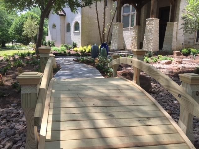 Wooden bridge over a dry creek bed leading to a house with landscaping.