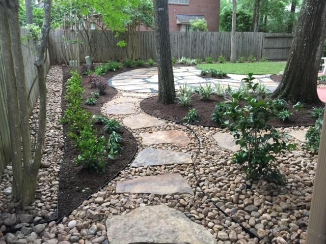 Stone path winds through a garden with trees, plants, and mulch, leading to a grassy area.