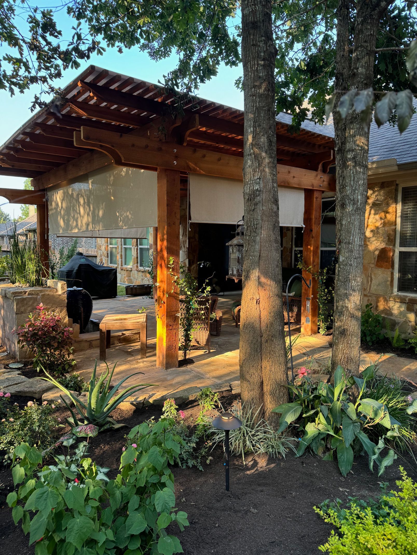 Wooden pergola over a patio, framed by trees and surrounded by a garden with plants.