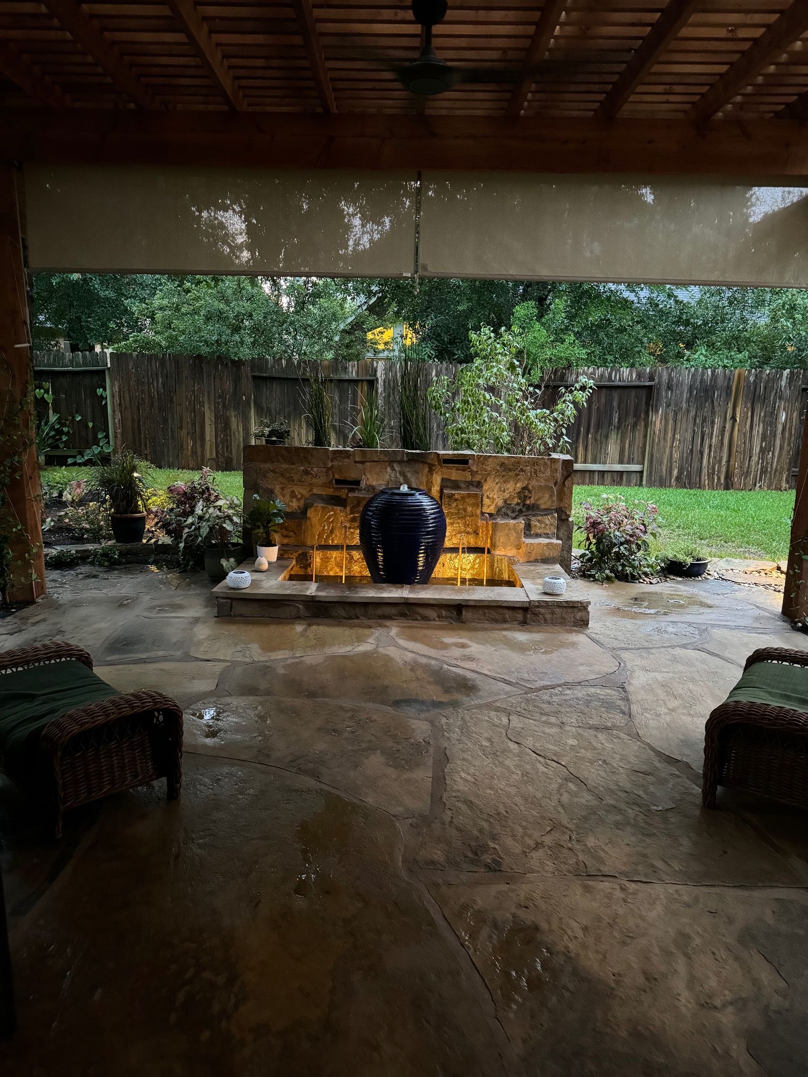 Covered patio with a dark blue fountain, stone wall, and lush greenery in the background. Rain is falling.