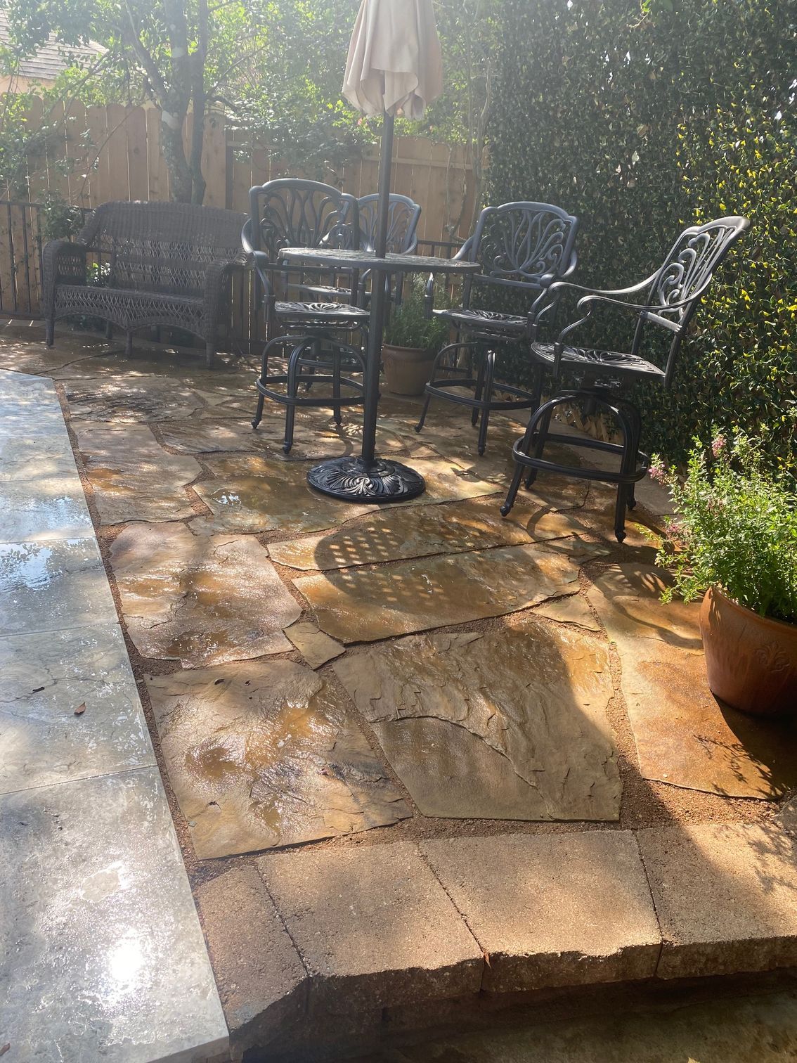 Patio with black wrought-iron furniture. Round table with chairs, bench, and umbrella. Stone walkway, green foliage.
