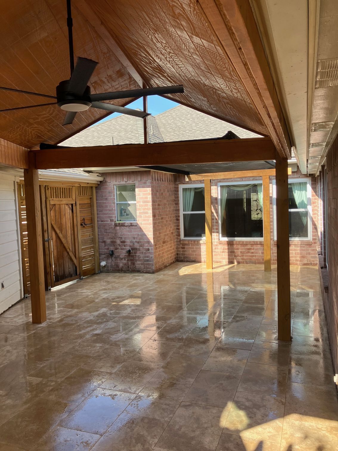 Covered outdoor patio with wood ceiling and supports. A ceiling fan hangs. Brick building visible.