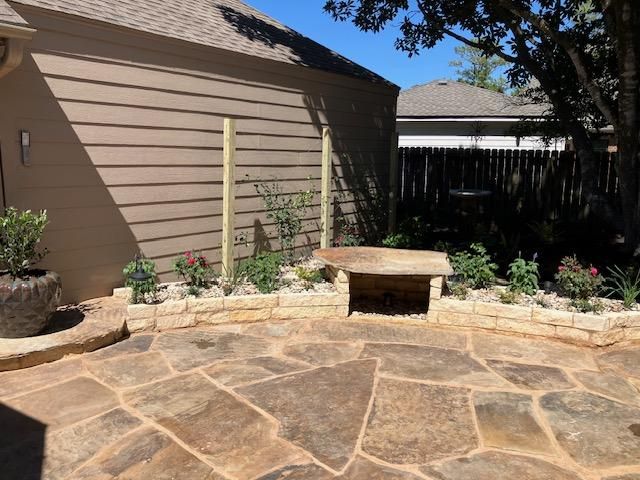 Stone patio with a stone planter and built-in bench. Flowers and a house with brown siding are visible.