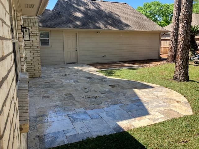 Stone patio with curved edge, adjacent to a beige house, lawn, and trees.