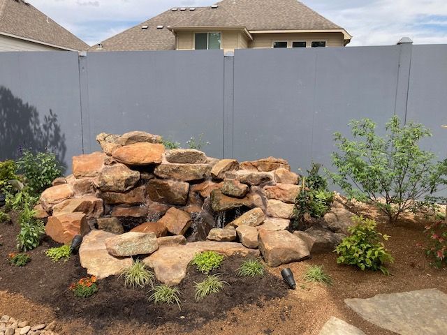 Rock waterfall feature in a yard with a grey fence and surrounding landscaping.