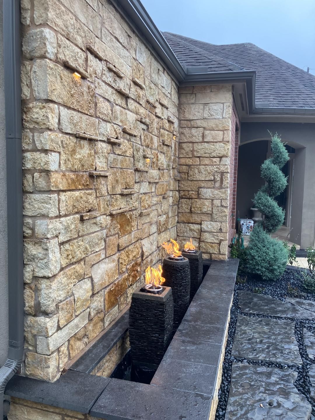 Stone water feature with flames, set against a stone wall and near a house.