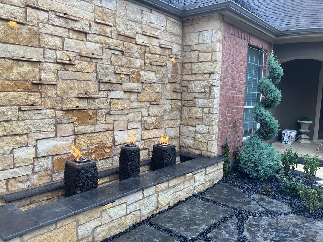 A stone water feature with flames, set in a yard, with a spiral topiary and dark paving stones.