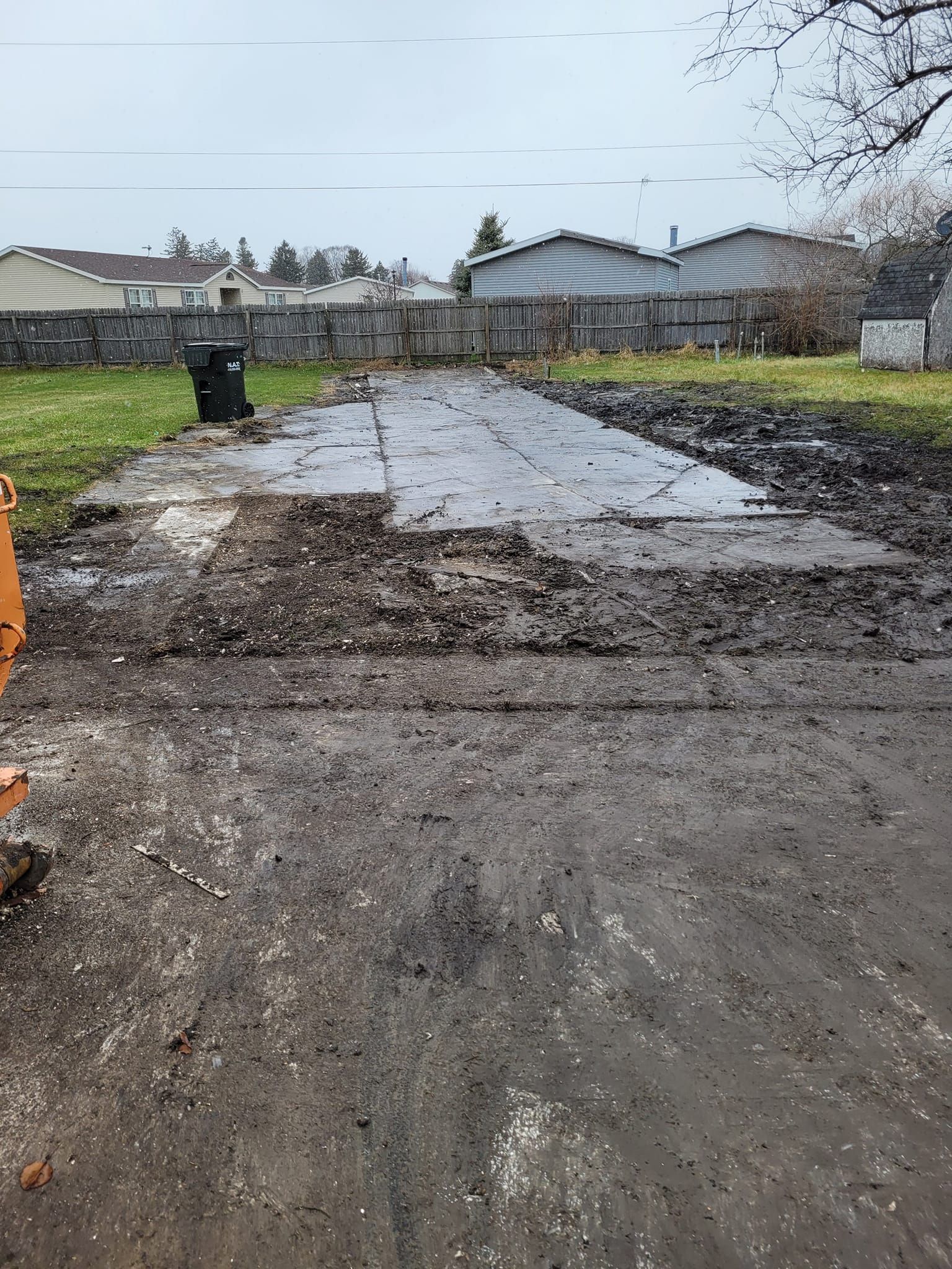Muddy driveway, cracked pavement, trash can. Backyard setting. Overcast sky.