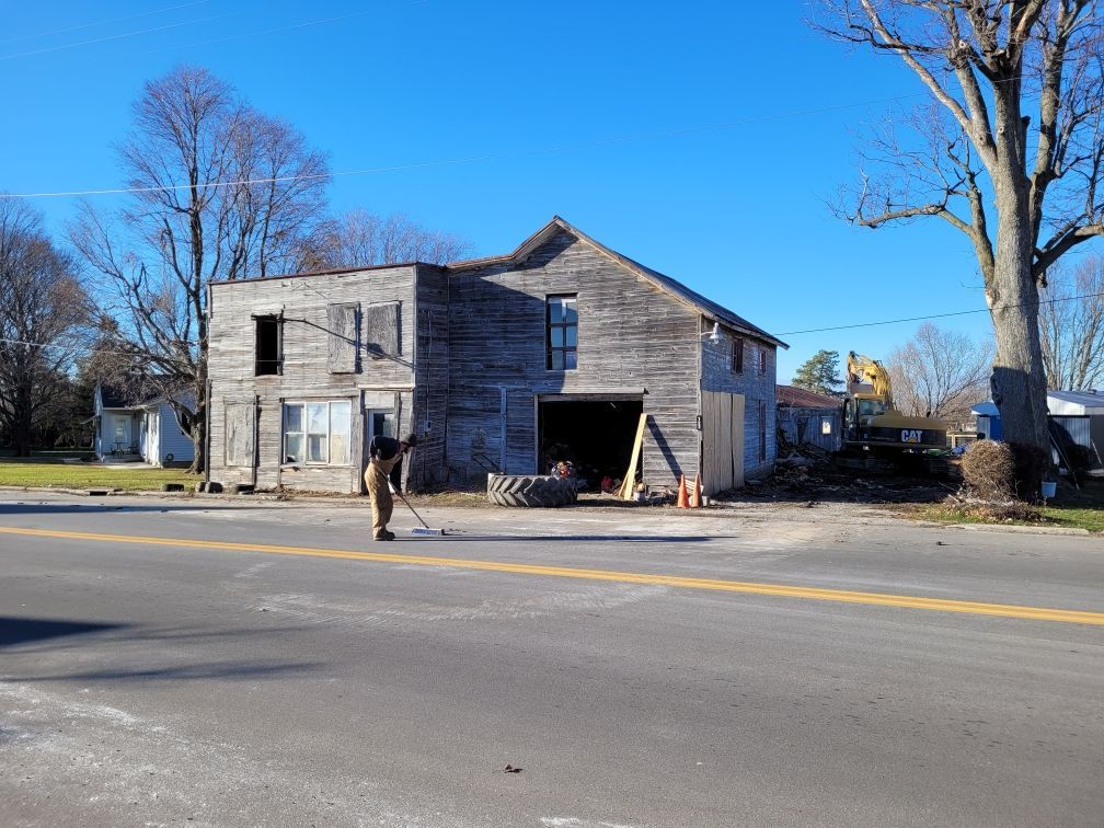Weathered building undergoing demolition next to a road on a sunny day; a worker sweeps.