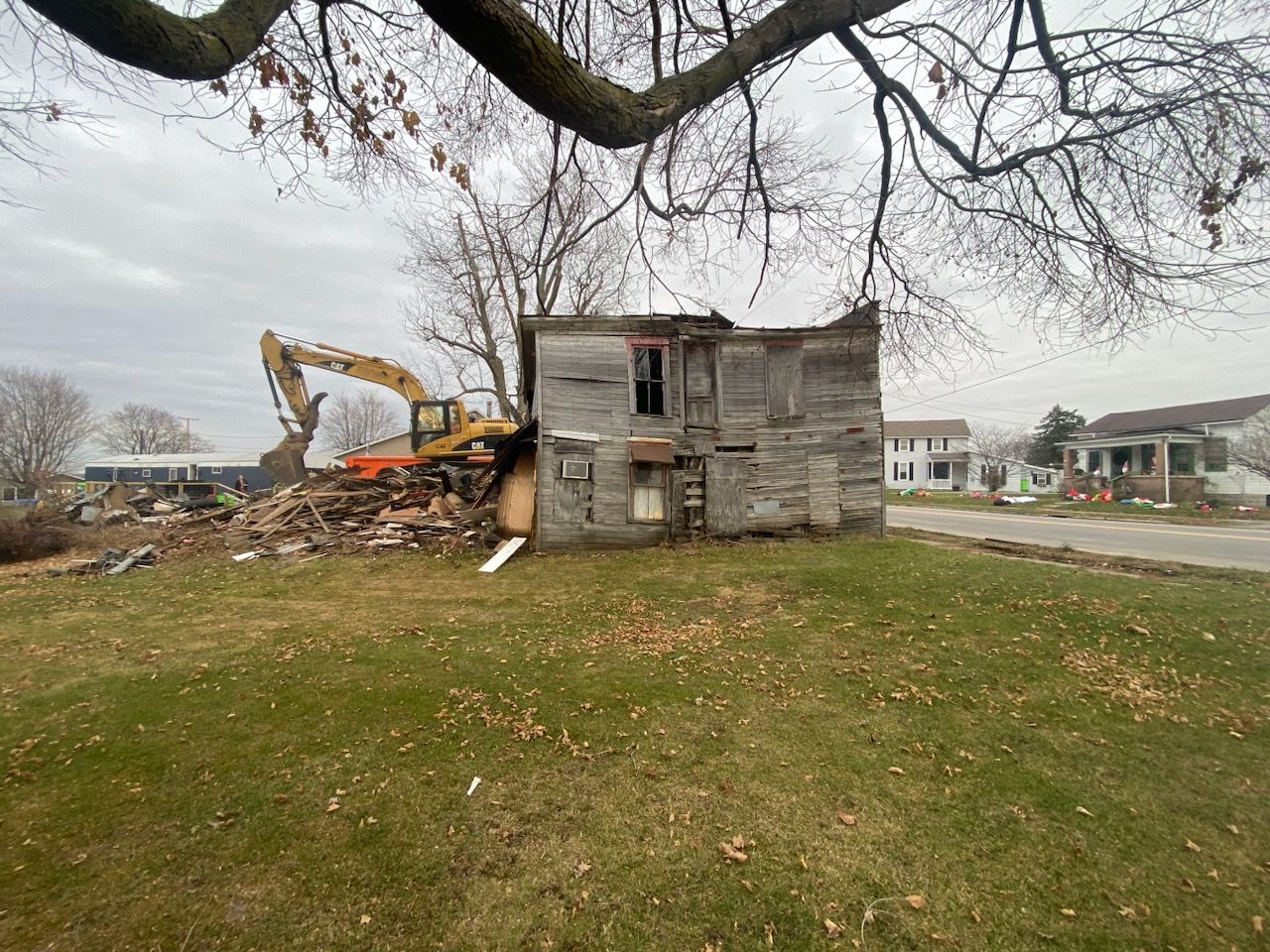 Demolition of a weathered wooden house, excavator at work. Grassy yard, overcast sky.