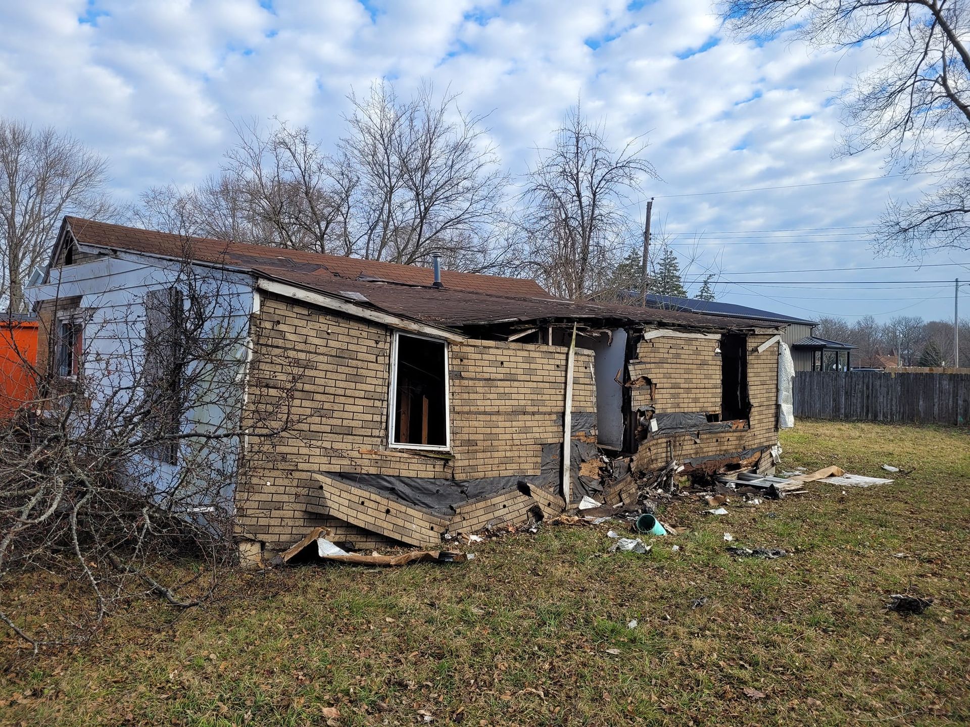 Damaged one-story brick house in disrepair, with missing walls and roof, surrounded by overgrown grass and bare trees.