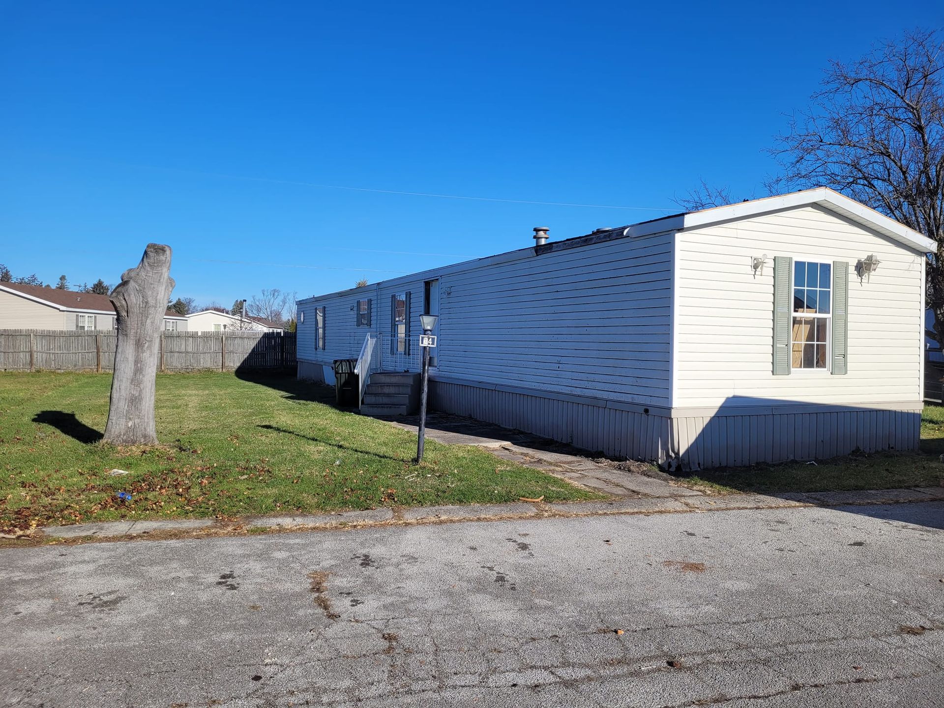White mobile home on a gravel lot under a clear blue sky. Green grass in front, dead tree on left.