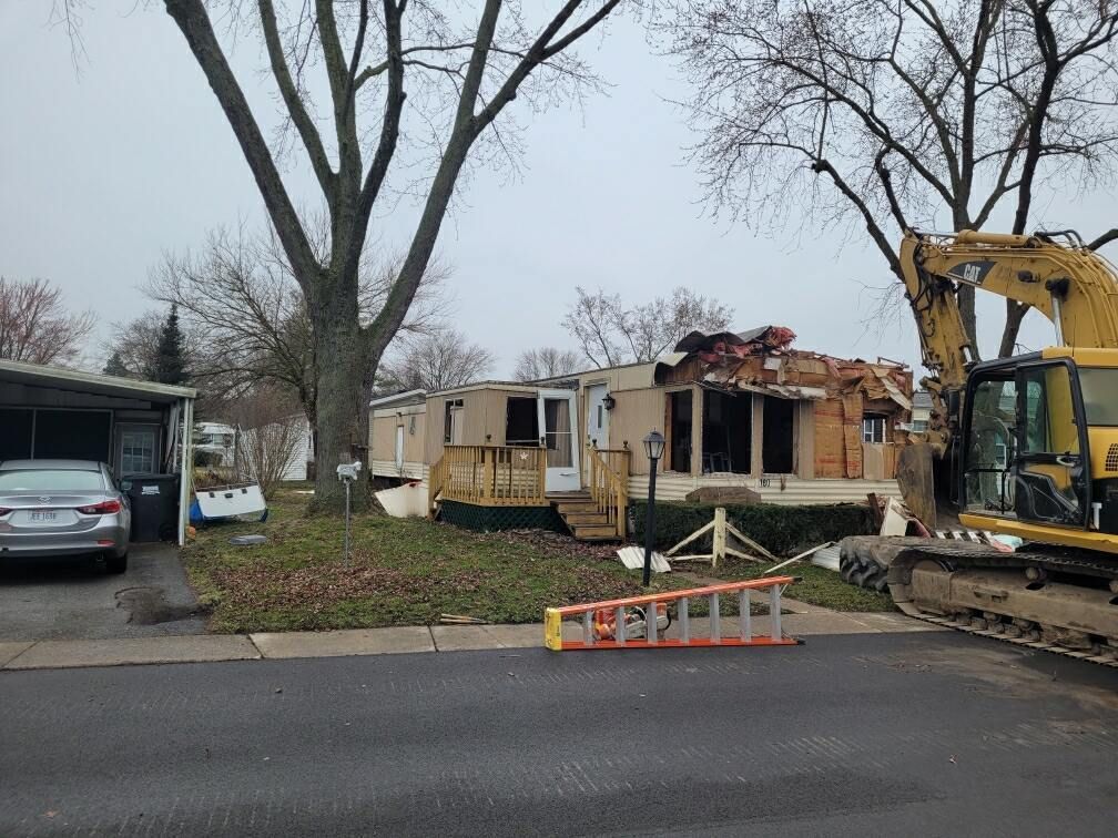 Mobile home partially demolished by excavator on a cloudy day.