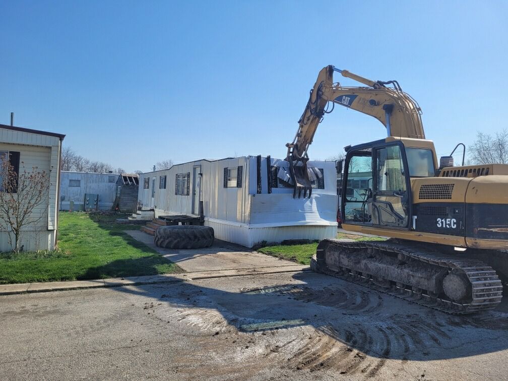 A yellow excavator demolishes a white mobile home on a sunny day.