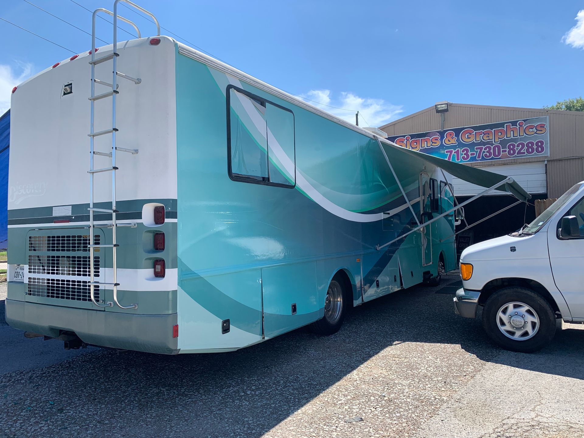 Rear view of a white and teal RV with a ladder. It's parked next to a white van and a building.