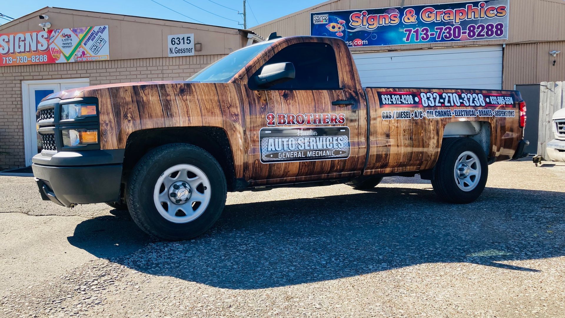 Brown camouflage-wrapped Chevrolet pickup truck parked outside a sign shop under a clear blue sky.