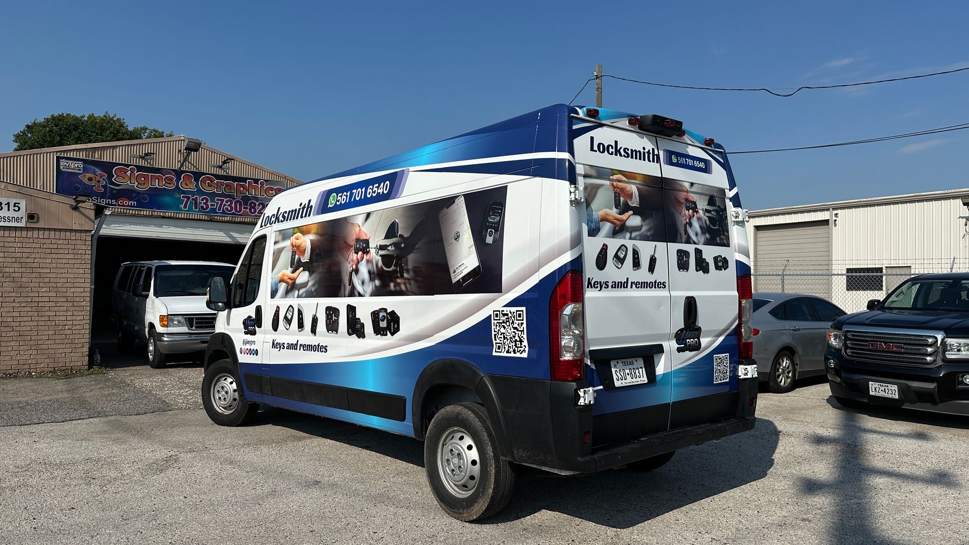 A blue and white locksmith van parked outside a garage, with a white vehicle inside and cars nearby.