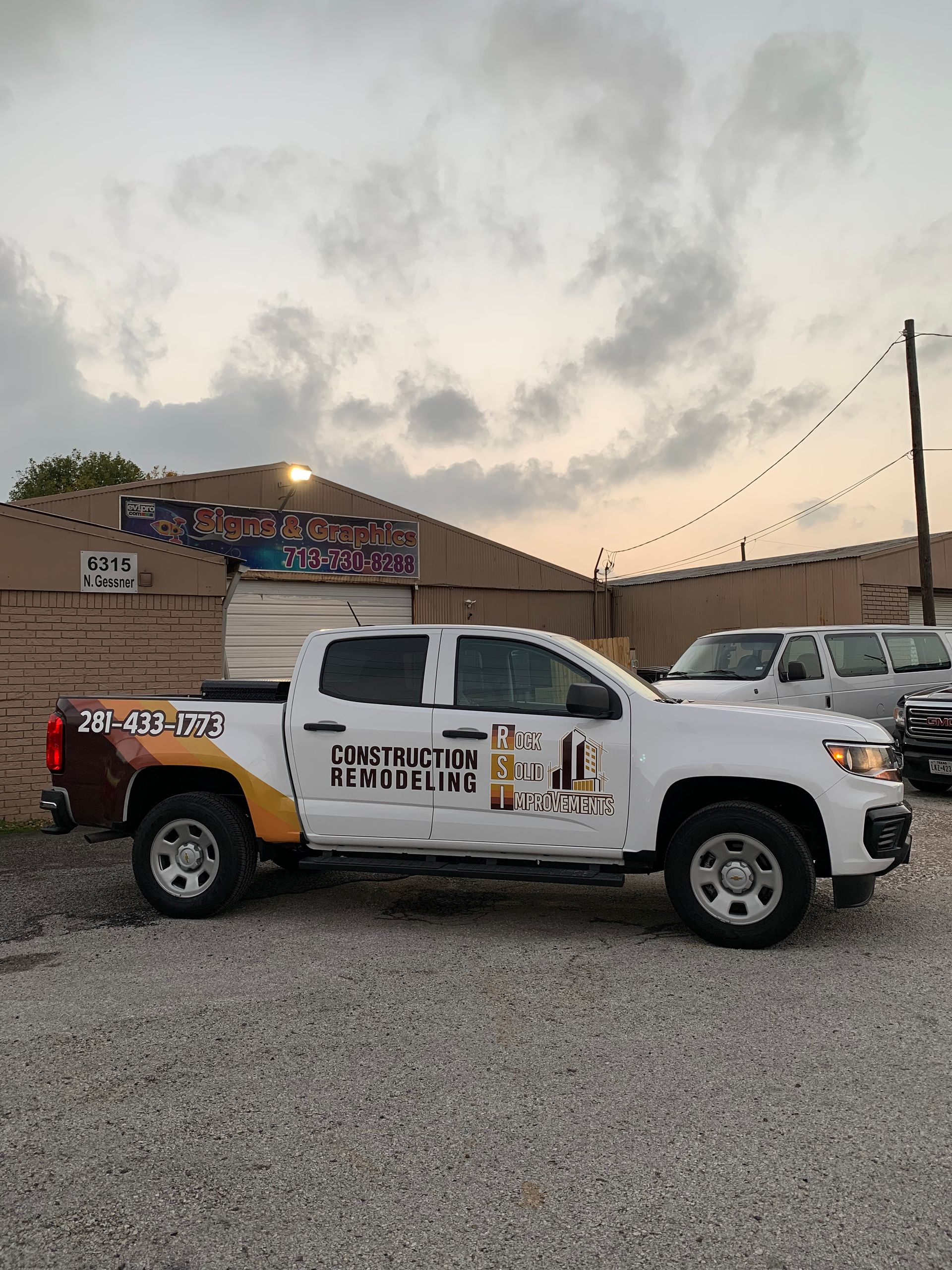 White construction truck parked, with company logo, in front of a building under a cloudy sky.