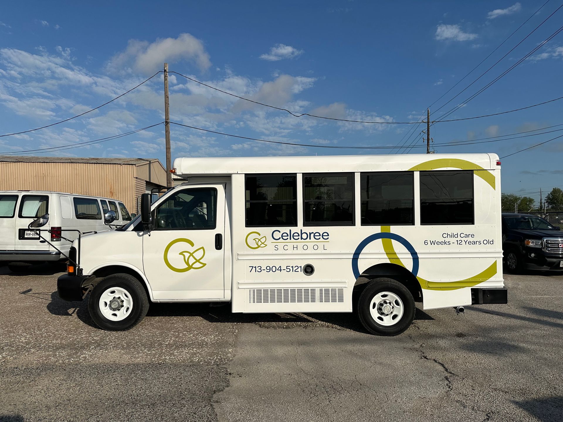 White bus with logo and phone number, parked outside with other vehicles on a cloudy day.