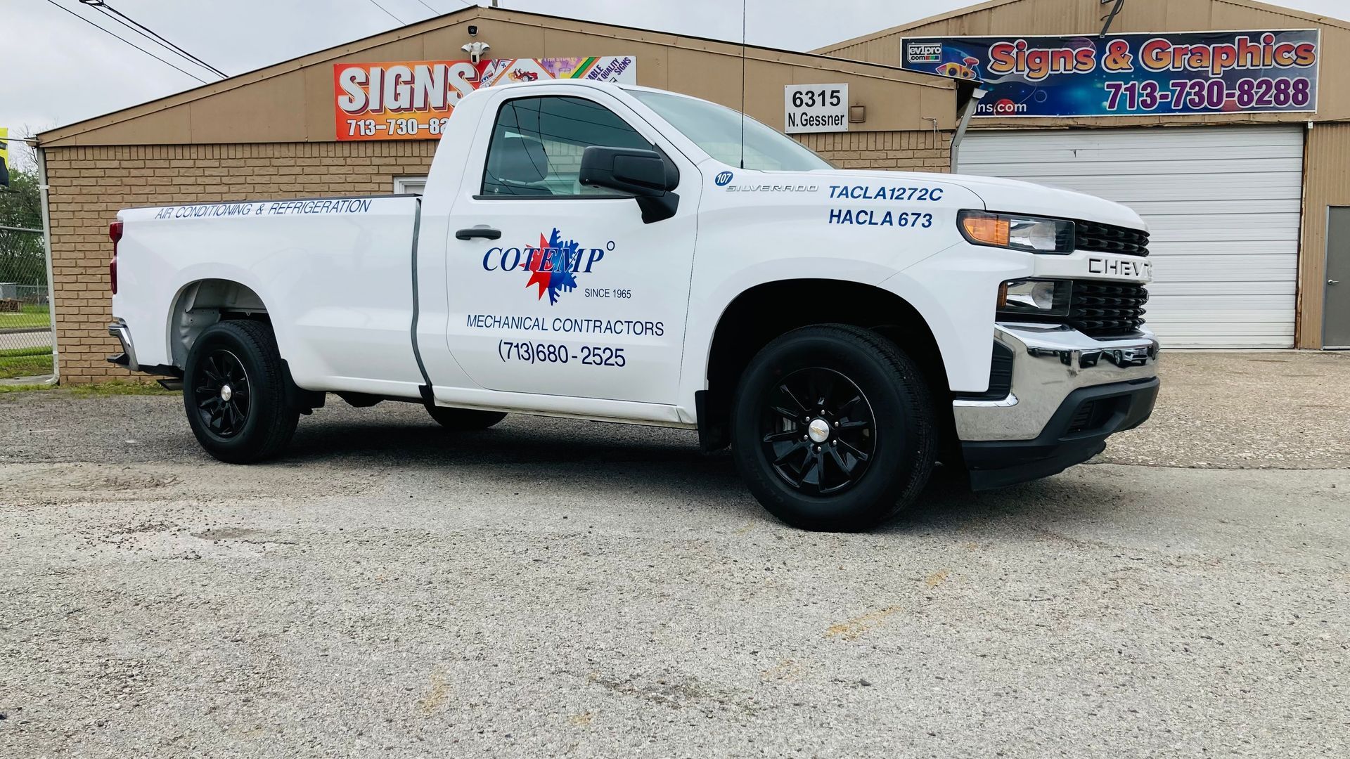 White Chevy pickup truck with black wheels parked in front of a sign shop.