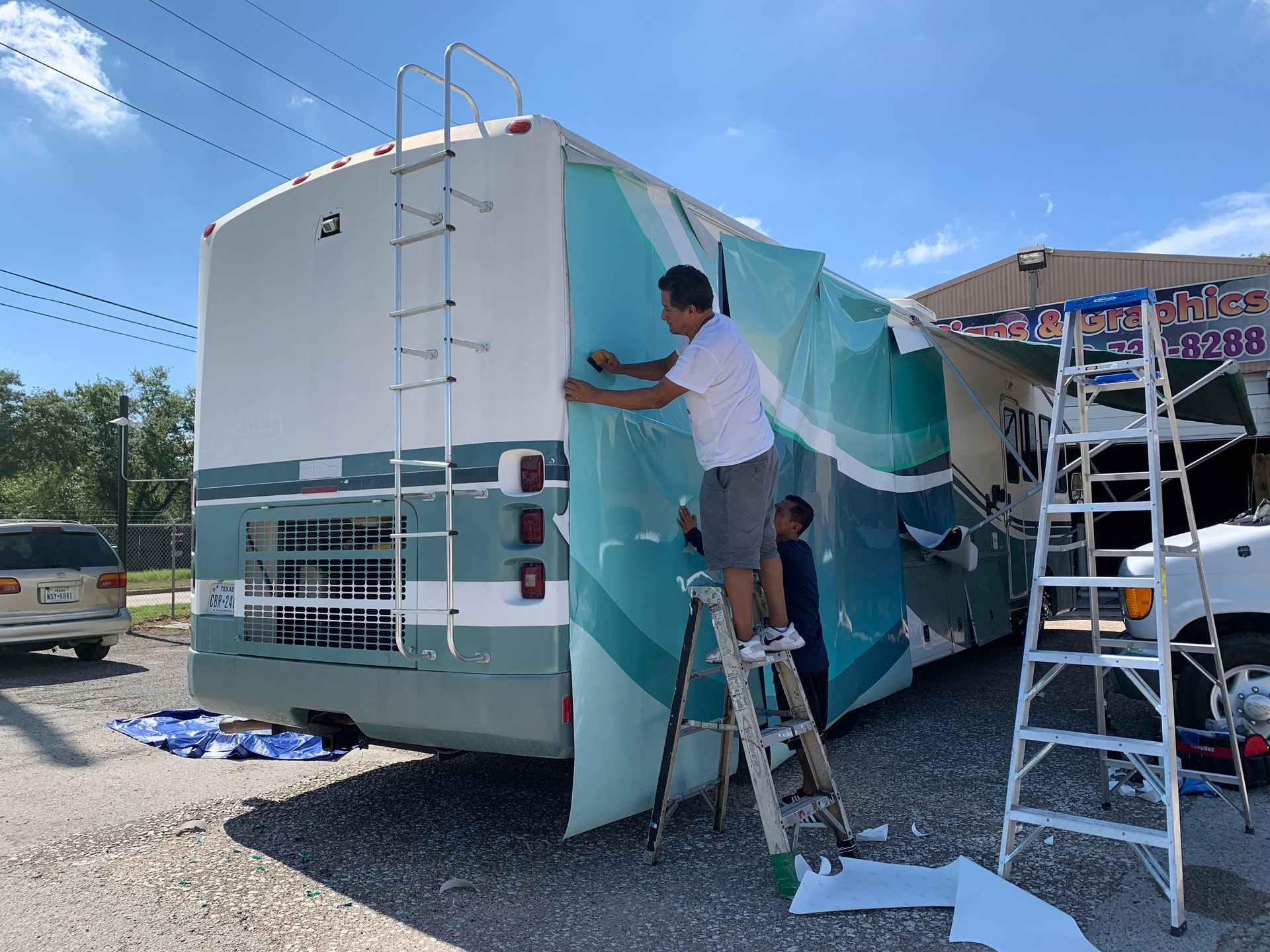 Two people applying a large teal and white graphic wrap to the side of a white RV, outdoors on a sunny day.