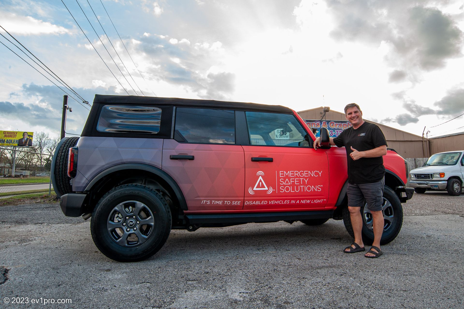 Man stands beside a red and purple Ford Bronco with logo, in a parking lot, thumbs up.