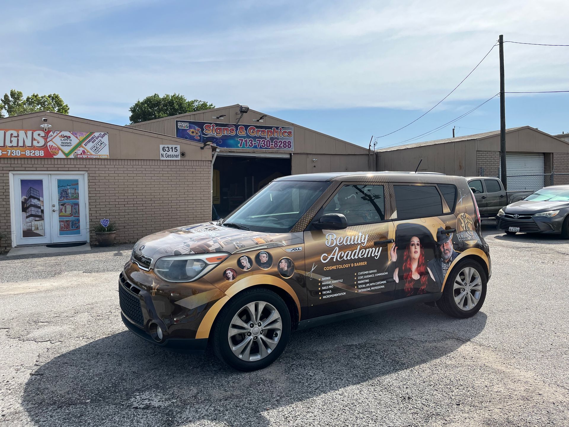Brown car with graphics in front of a sign shop; blue sky.