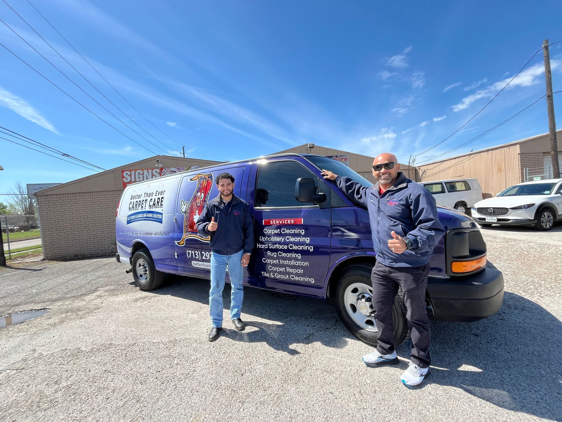 Two men stand beside a blue van with business signage. They both give a thumbs up, sunny day.