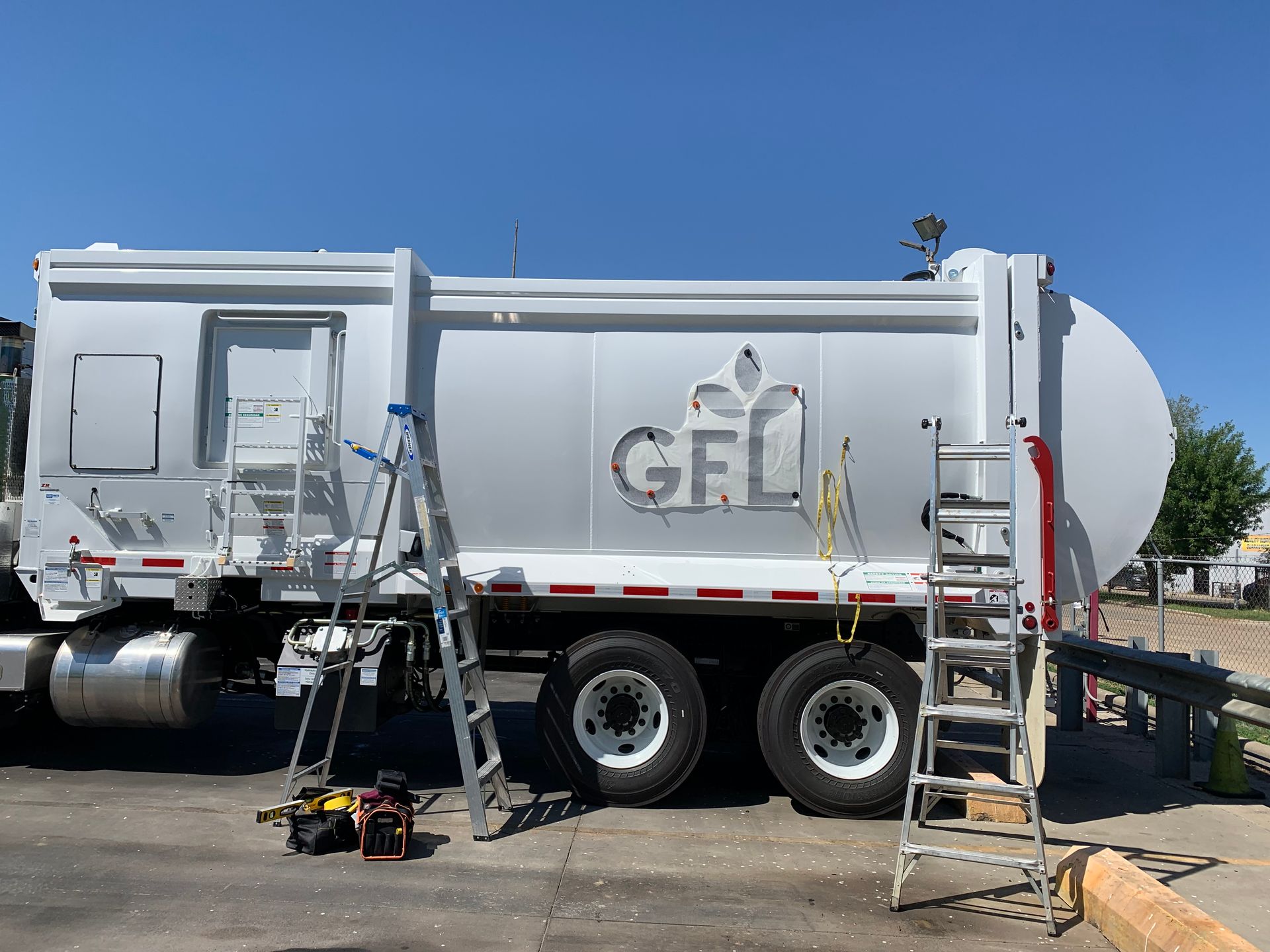 White GFL garbage truck with ladders and tools; parked in a lot, being worked on under a blue sky.