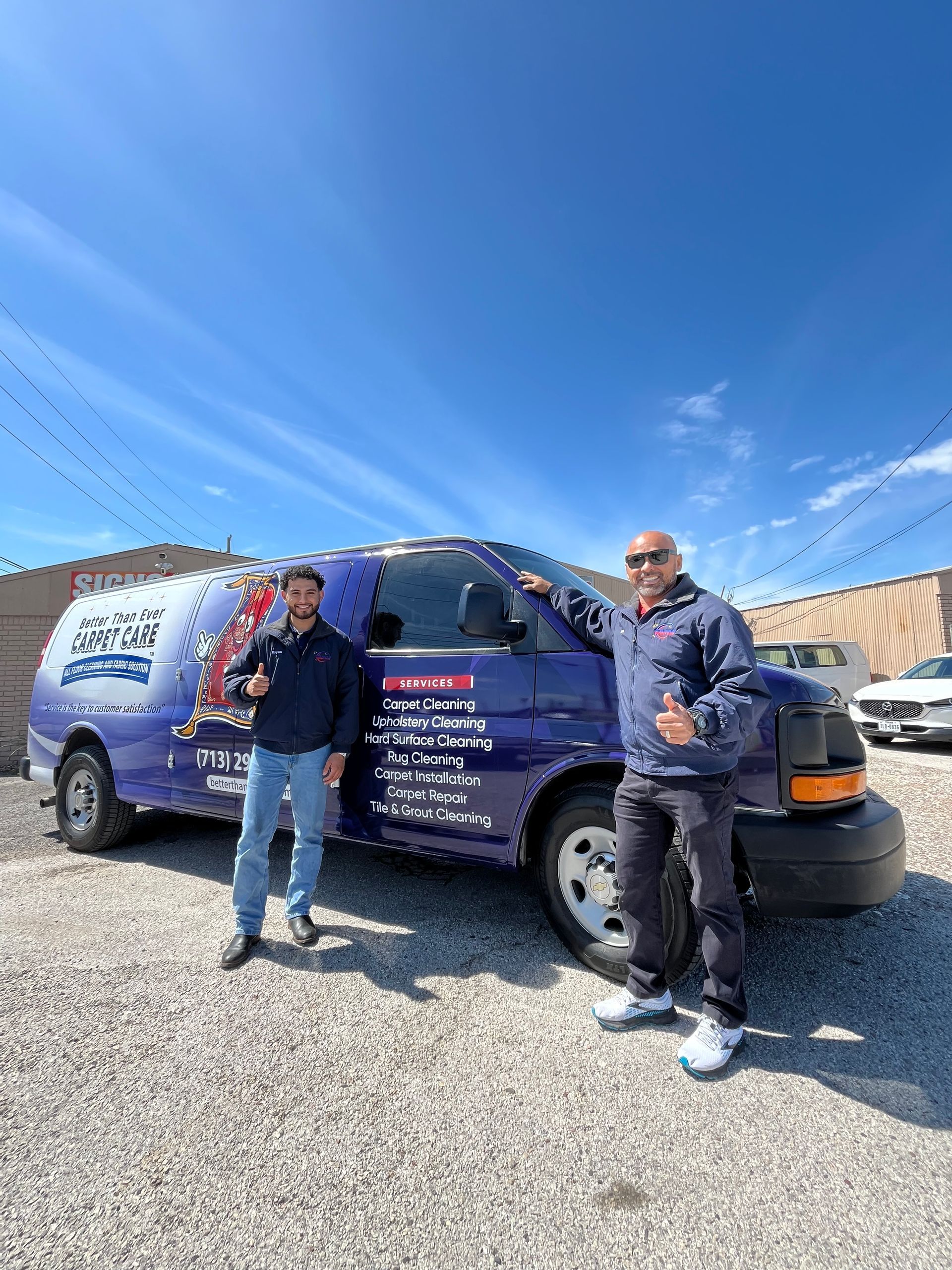 Two men stand next to a blue service van on a sunny day. They are giving thumbs up.