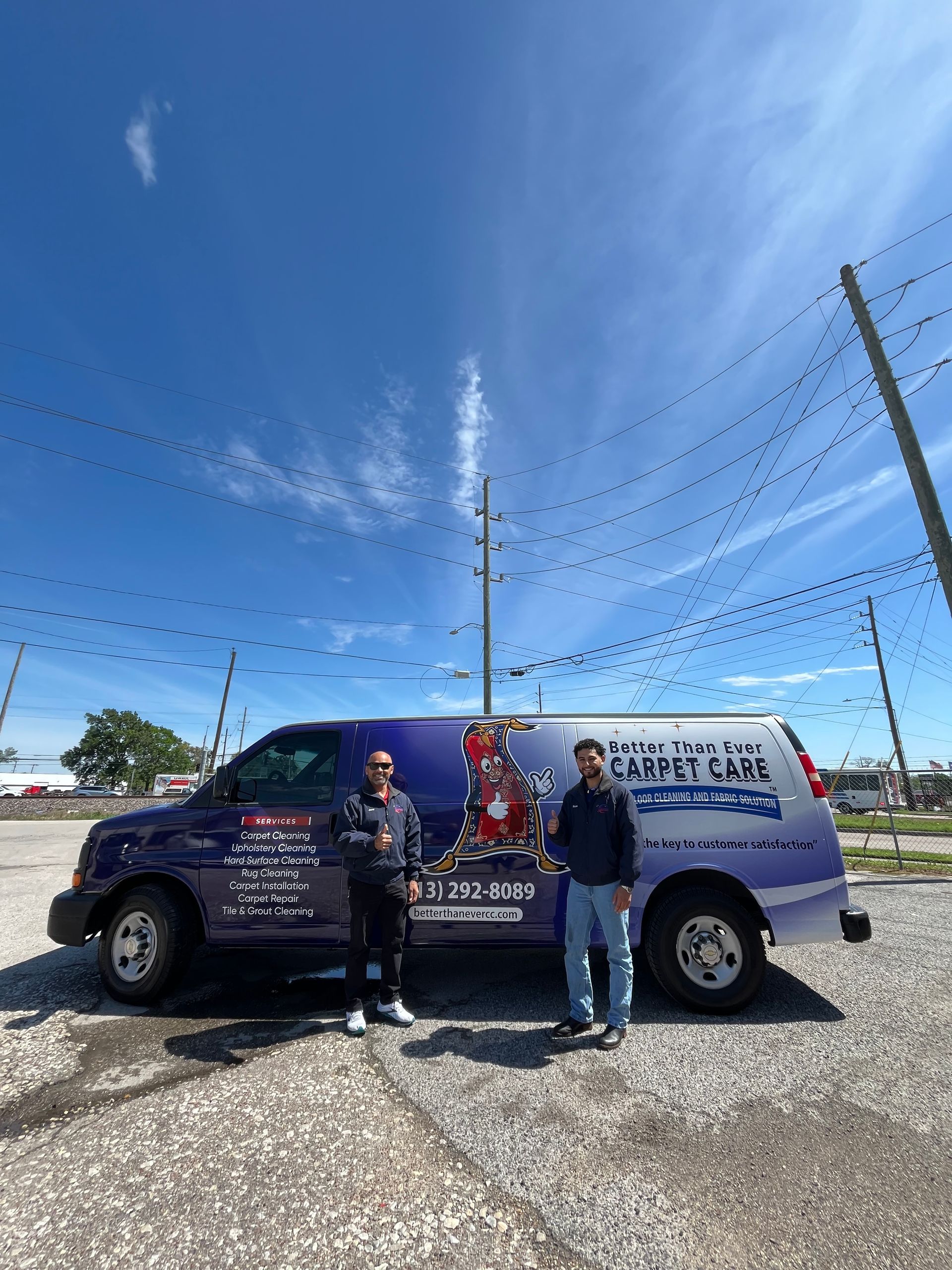 Two men stand by a blue company van in a parking lot on a sunny day. The van has text and logos.