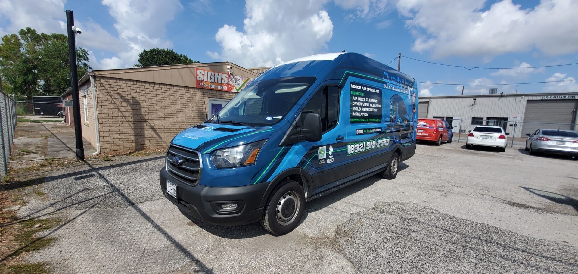 Blue van parked in a gravel lot with buildings and sky in the background.