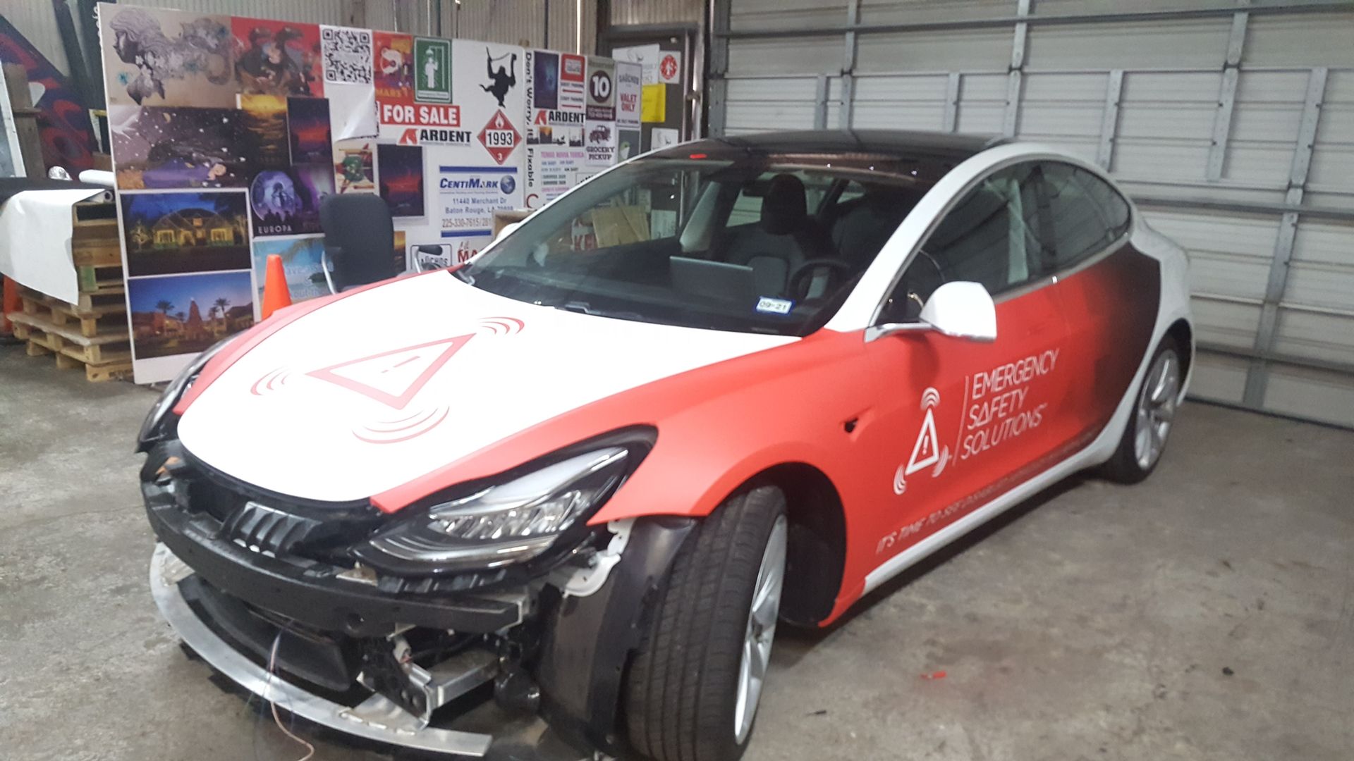 Damaged white and red Tesla Model 3 in a garage, front bumper missing.