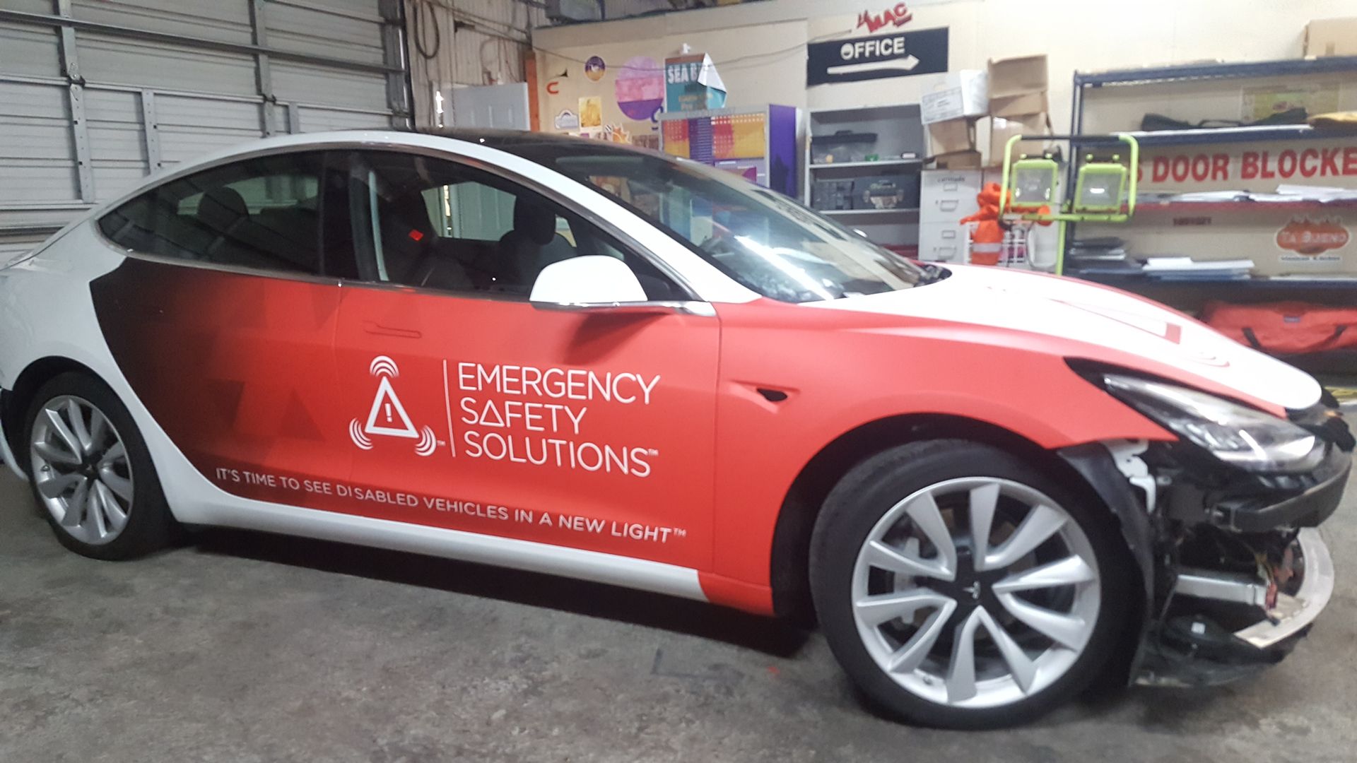 Tesla car with red and white Emergency Safety Solutions wrap, in garage with front bumper removed.