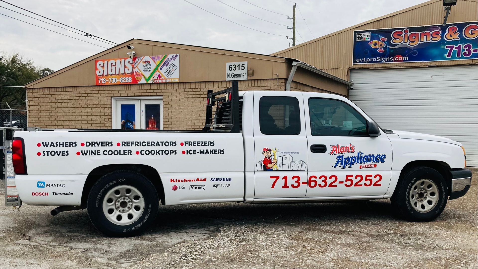 White work truck parked in front of a building with business signage. Truck has company logos and a phone number.