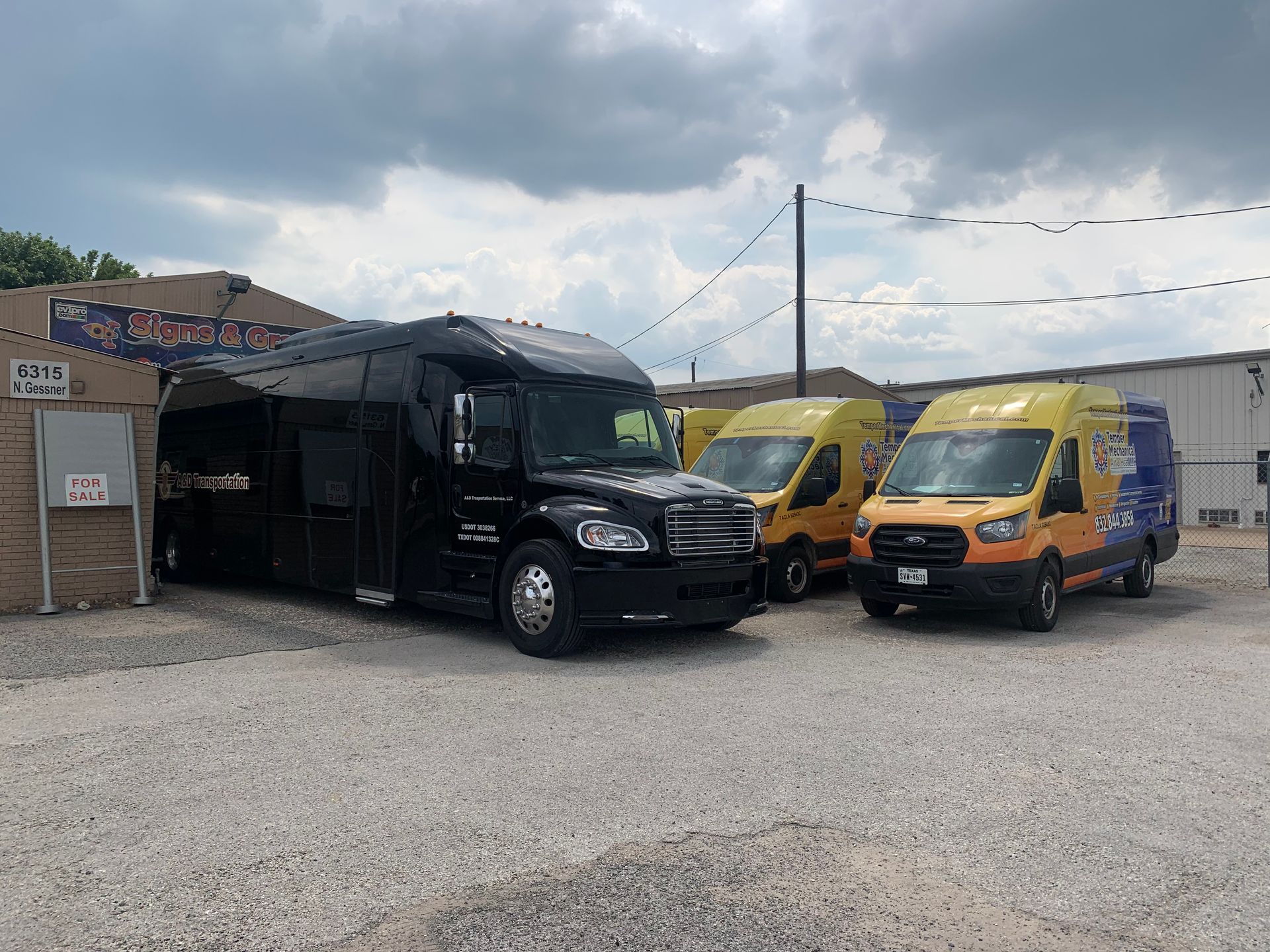Black bus and two yellow vans parked outside a business under a cloudy sky.