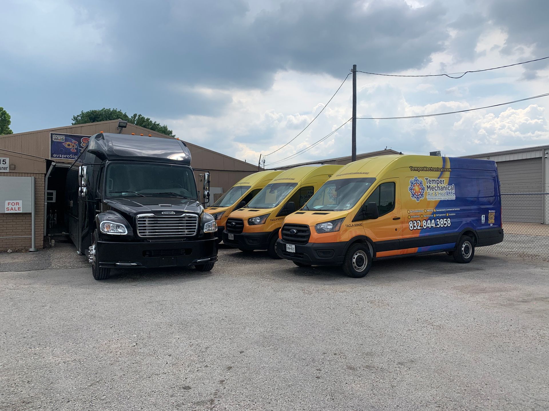 Black truck and three yellow vans parked in front of a building on a gravel lot under a cloudy sky.